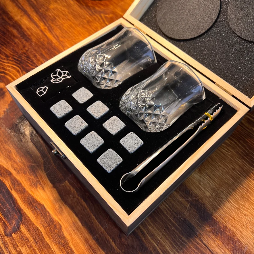 Caja de madera abierta con dos vasos de cristal, ocho piedras para enfriar bebidas y unas pinzas metálicas sobre un forro negro