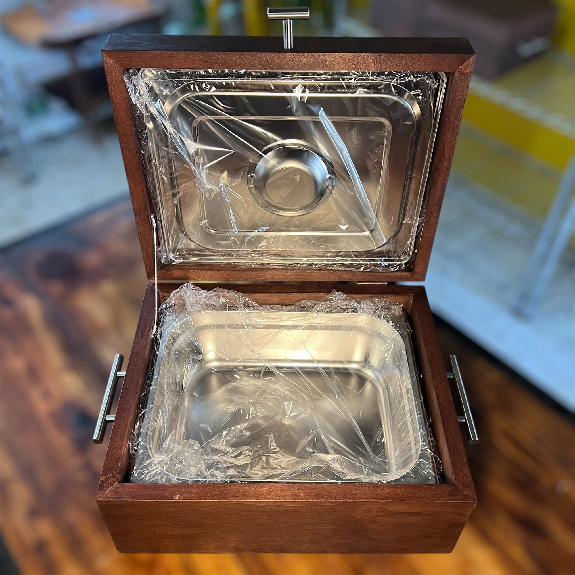 Caja de madera oscura abierta con dos recipientes de acero inoxidable envueltos en plástico transparente