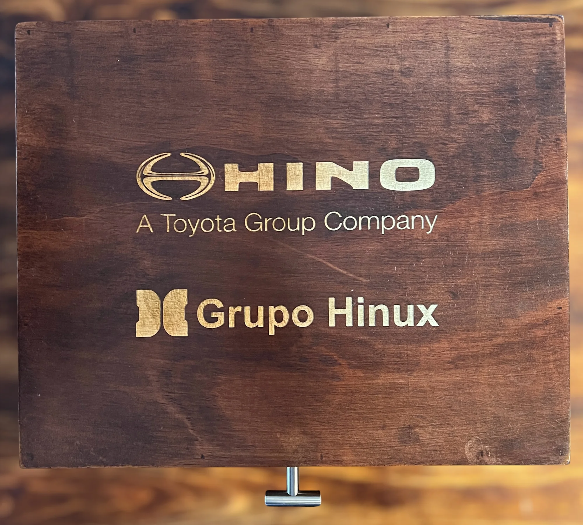 Tapa de madera oscura con los logotipos dorados de Hino y Grupo Hinux, y un asa metálica plateada en la parte inferior