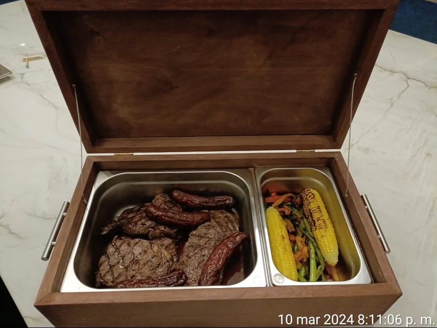 Caja de madera oscura abierta con dos recipientes de acero inoxidable llenos de carne asada y elotes con verduras