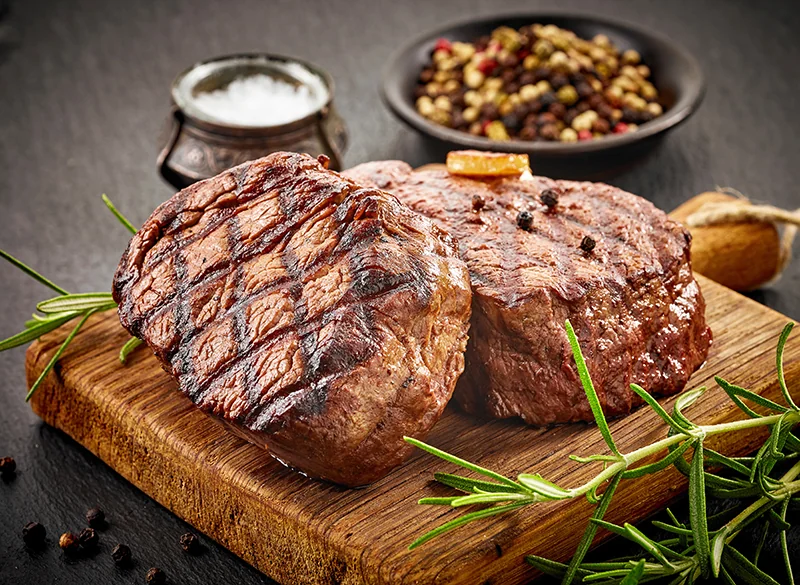 Dos cortes de carne asada con marcas de parrilla, romero, granos de pimienta y sal en una tabla de madera