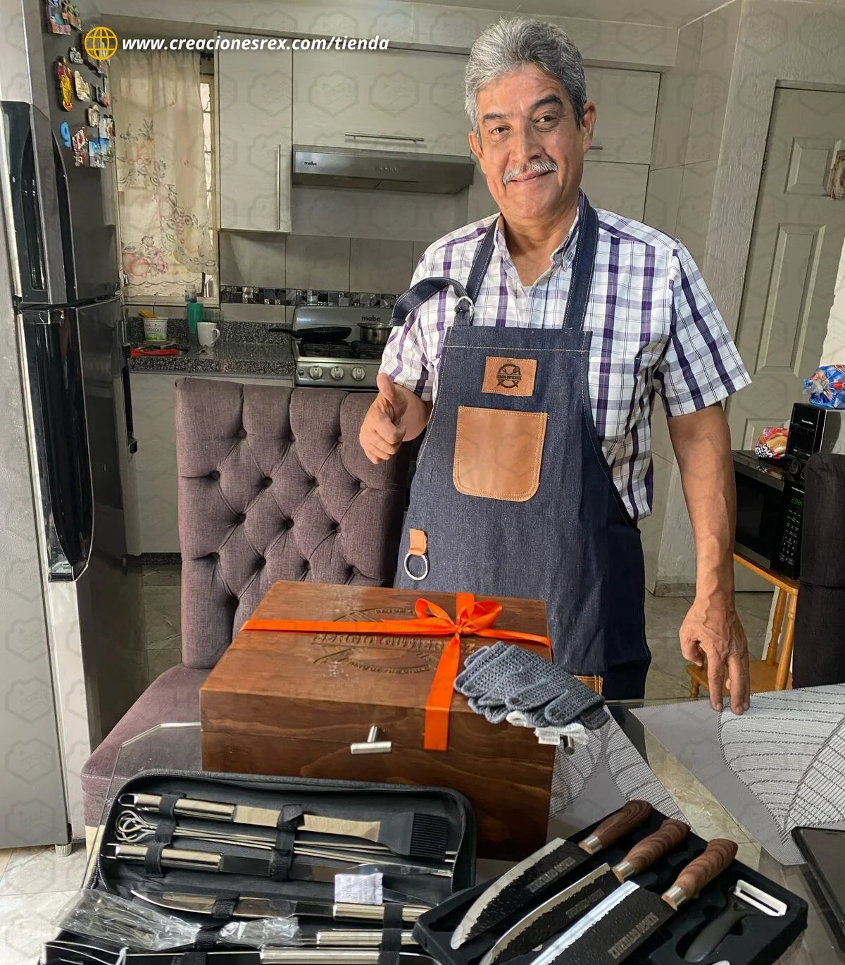 Hombre sonriente con delantal de mezclilla y un pulgar arriba, junto a una caja de madera con un moño naranja, guantes y estuches de cuchillos y utensilios para parrilla
