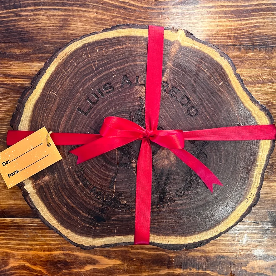 Tabla de madera oscura con forma irregular, grabada con el nombre "Luis Alfredo", atada con un listón rojo y una etiqueta de regalo
