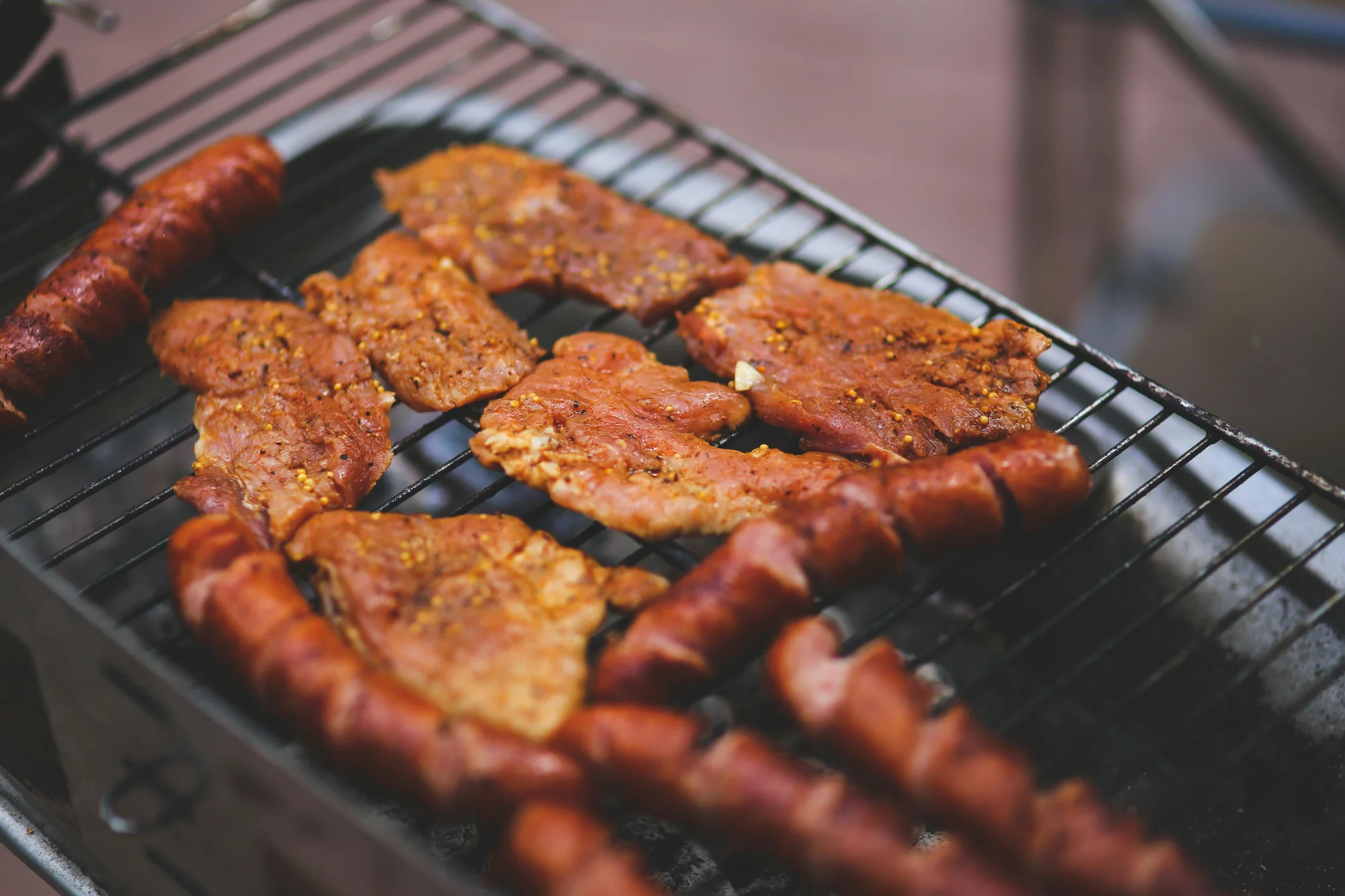 Trozos de carne marinada y salchichas asándose en una parrilla de metal, con un fondo oscuro y desenfocado