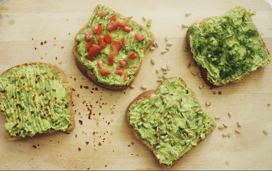 Cuatro tostadas de aguacate con diferentes toppings: tomate, semillas de girasol, cilantro y hojuelas de chile
