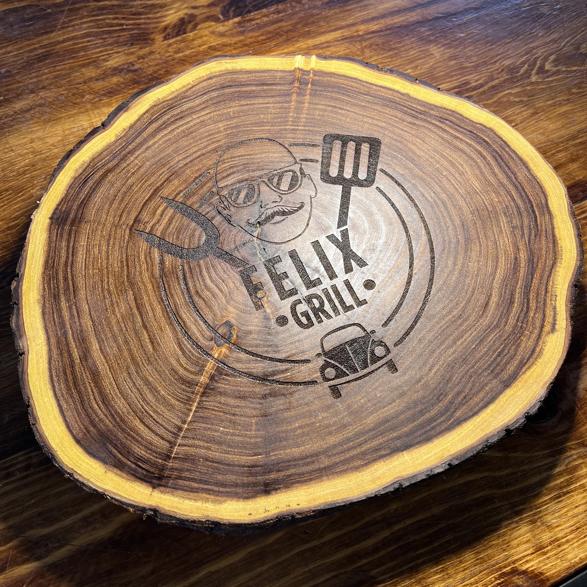 Tabla de madera rústica con corteza amarilla y grabado láser de un hombre con lentes, utensilios de parrilla y un auto, sobre una mesa de madera