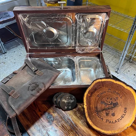 Caja de madera abierta con recipientes de acero inoxidable, un mandil de cuero, posavasos y una tabla de madera con grabado de un oso parrillero