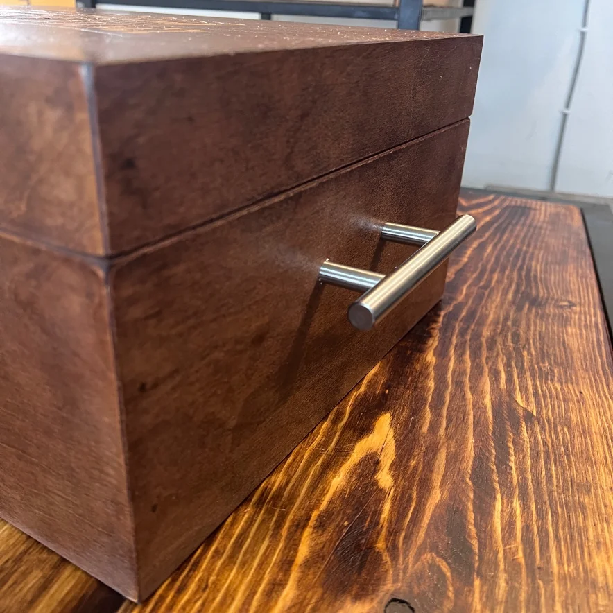 Caja de madera oscura con una manija metálica plateada, sobre una superficie de madera con vetas marcadas