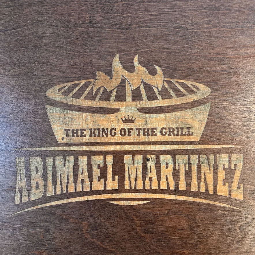 Grabado láser en madera oscura con un diseño de parrilla, llamas y una corona, con el texto