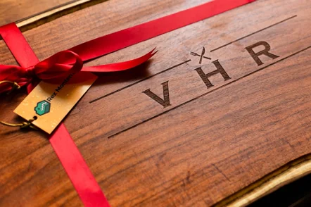Tabla de madera con un moño rojo y una etiqueta, y las iniciales VHR grabadas con un cuchillo y un tenedor