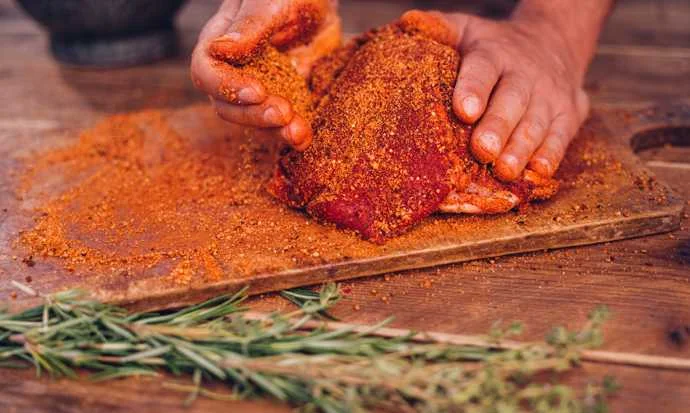 Manos espolvoreando un adobo naranja sobre un corte de carne roja en una tabla de madera, con hierbas frescas al frente