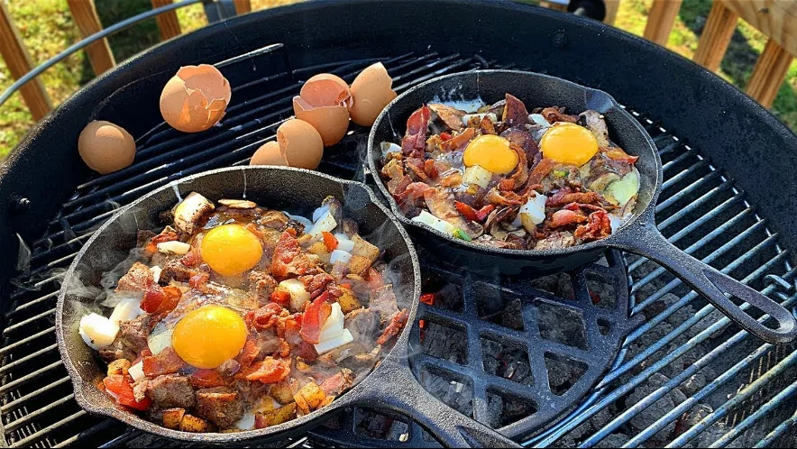 Dos sartenes de hierro fundido con huevos, tocino y papas sobre la parrilla de un asador, con cáscaras de huevo a un lado
