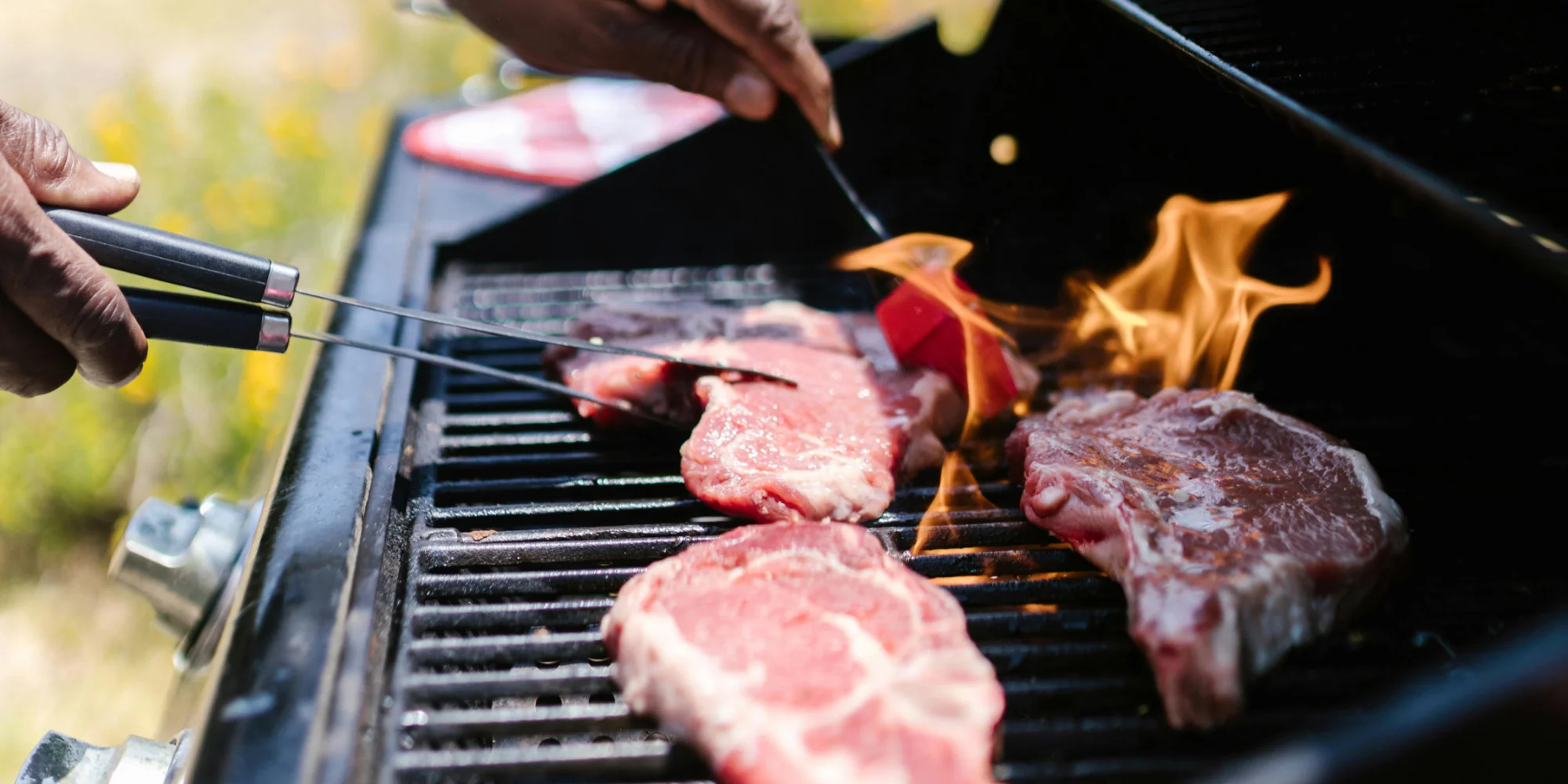 Manos volteando cortes de carne cruda sobre una parrilla con fuego y llamas, mostrando el proceso de cocción