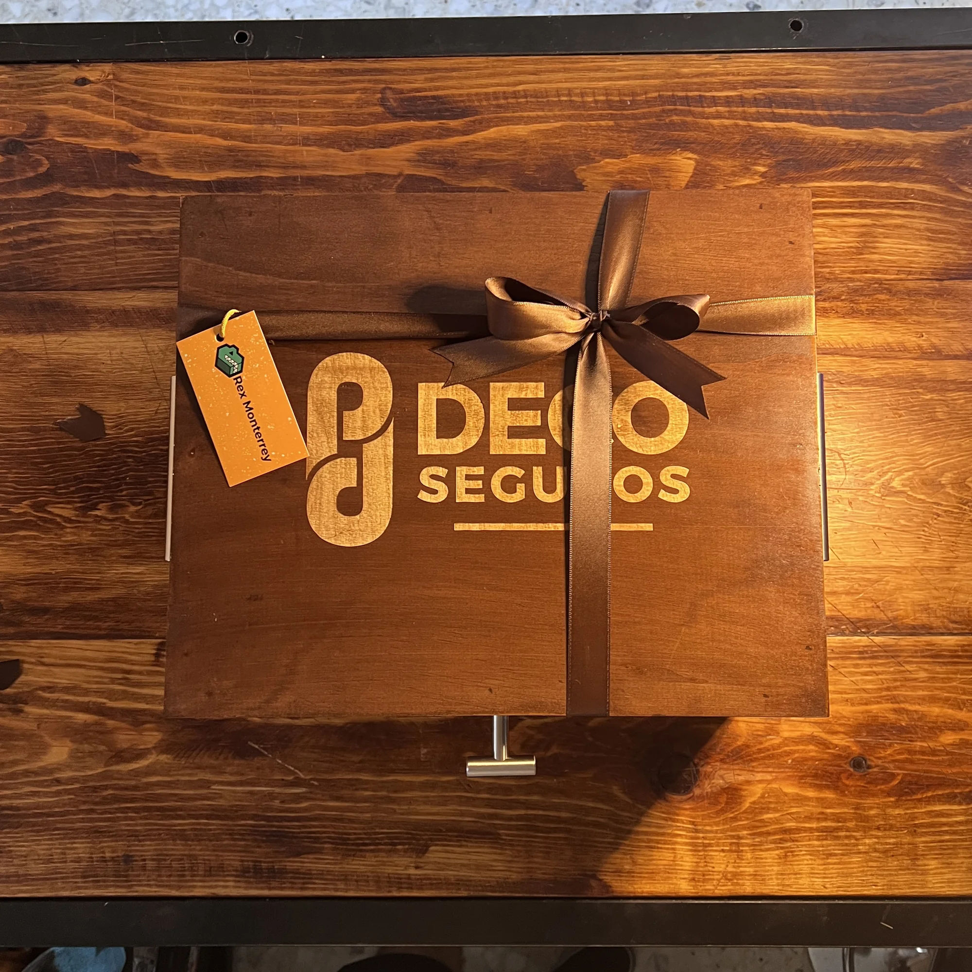 Caja de madera oscura con un moño café, un logo que dice "DECO SEGUROS" y una etiqueta naranja de