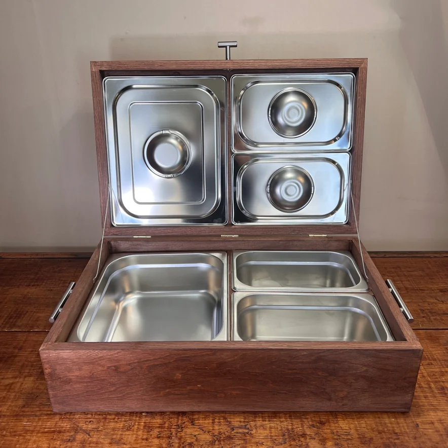 Caja de madera oscura abierta con cuatro recipientes de acero inoxidable, dos en la tapa y dos en la base