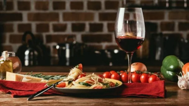 Plato de pasta con tomates cherry y albahaca, un vaso de vino tinto, y vegetales frescos sobre una mesa de madera