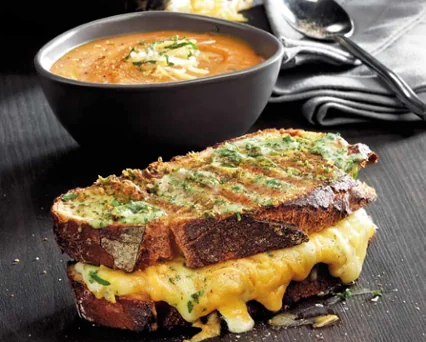 Sándwich de pan tostado con queso derretido y perejil, acompañado de un tazón de sopa de tomate y una cuchara