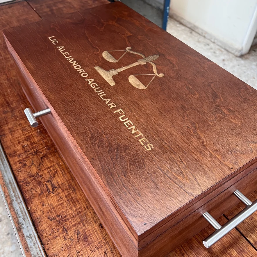 Caja de madera oscura con grabado láser de una balanza y el nombre