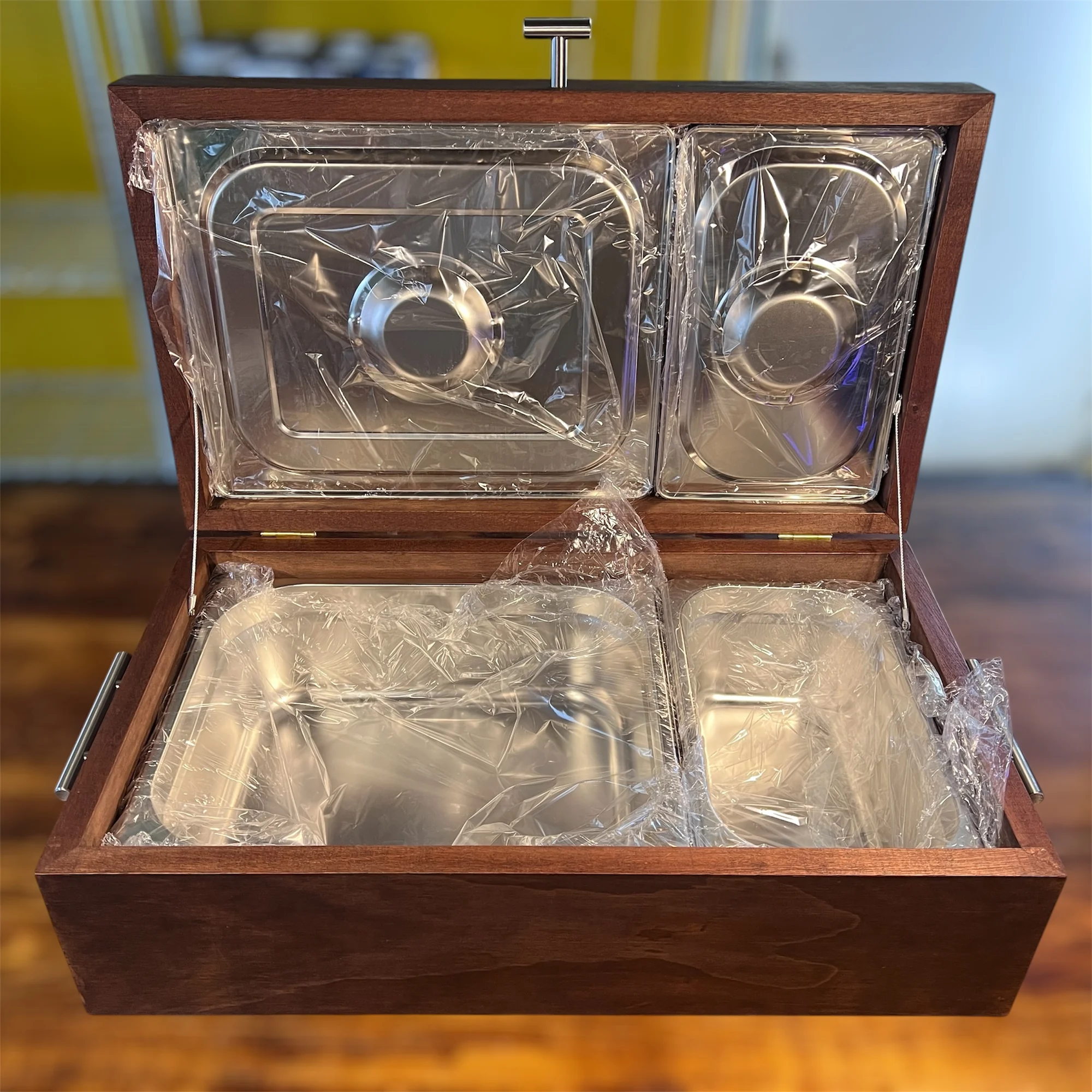 Caja de madera oscura abierta con cuatro recipientes de acero inoxidable cubiertos con plástico transparente