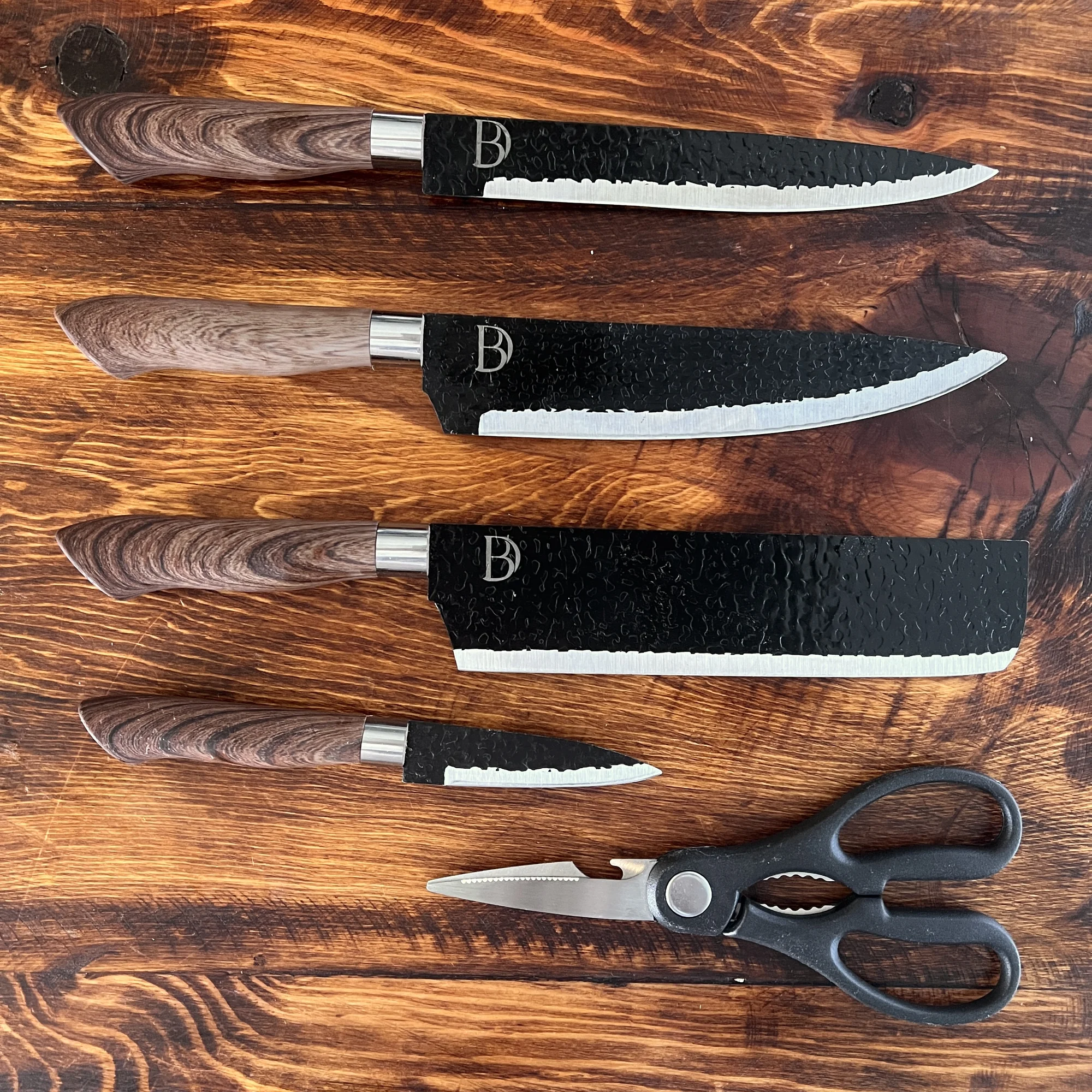 Juego de cuatro cuchillos con mangos de madera y hojas negras texturizadas, y unas tijeras de cocina, sobre una tabla de madera