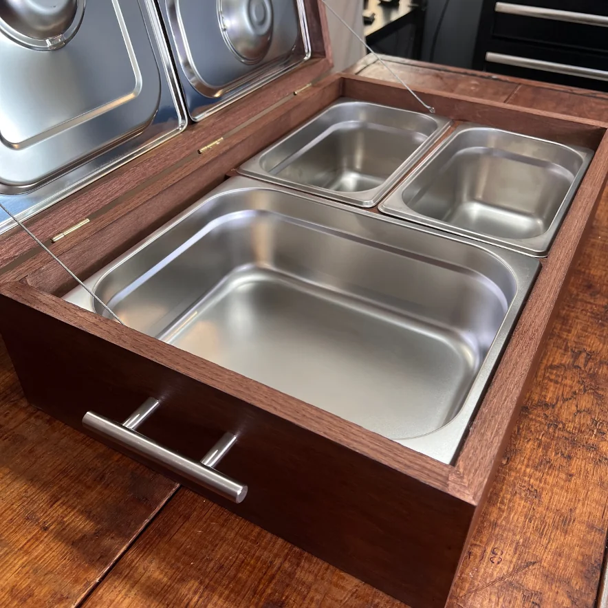 Caja de madera oscura con tres recipientes de acero inoxidable y una tapa abierta con dos recipientes más